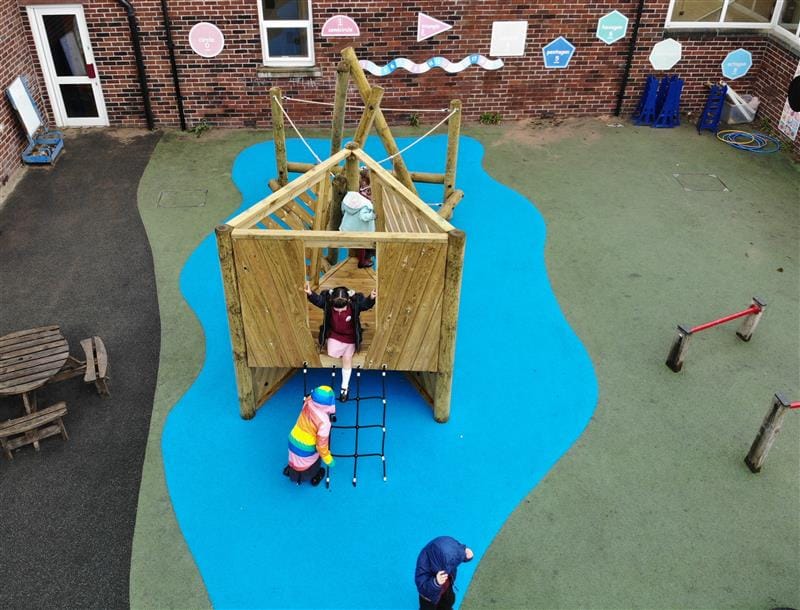 3 children, one wearing a multi-coloured raincoat are playing on a climbing frame that has been installed onto blue wetpour surfacing in front of the school building. 