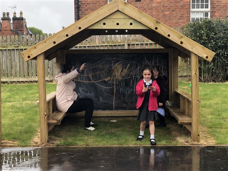 Two children are stood in the Giant Playhouse as they scribble on the chalkboard attached to the back wall. A teacher is sat inside to, talking to both of the children and encouraging role play.