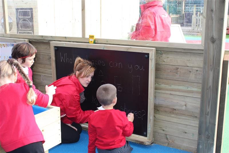 A little boy is holding a piece of chalk as he looks at a chalkboard. A teacher has knelt next to him and is teaching him what to write on the board. Two children can be seen behind the teacher as they watch the little boy start to write on the board.