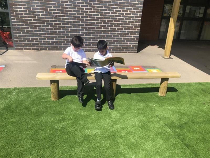 2 children wearing white polo t-shirts and black trousers sat on a bench reading books, the bench has been installed onto artificial grass in front of the school building. 