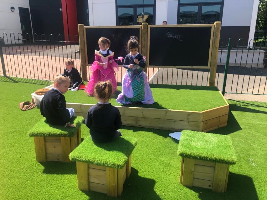 Two little girls are stood on a stage and are performing a play whilst a small group of children are watching from artificial grass topped seats.