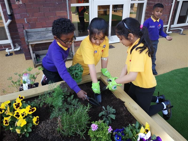 children stand around the corner of a planter wearing green gardening gloves and prodding the soil
