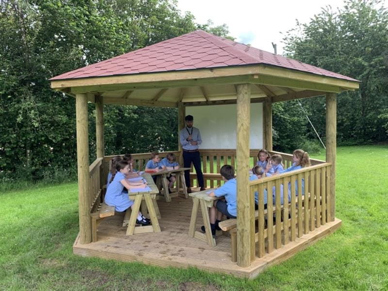 children sit in the outdoor classroom and have a lesson led by a teacher