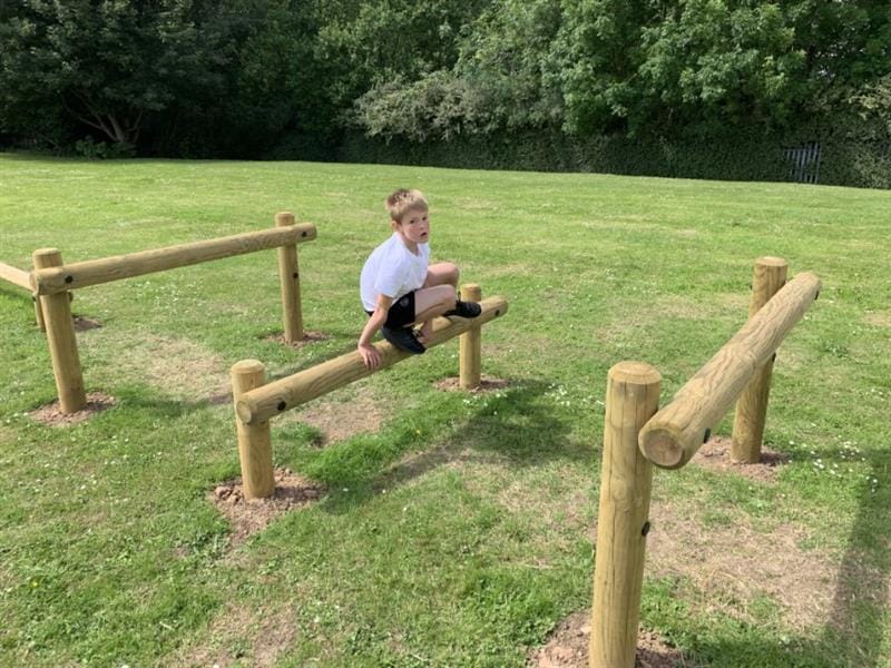 One boy wearing a white polo top is jumper over timber posts as part of an obstacle course. The timber posts have been installed into the grass. 