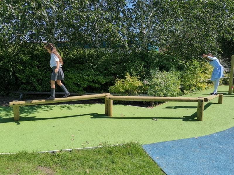 Two girls, one wearing a summer dress and one wearing a grey skirt and white top are playing on a trim trail that has been installed onto wetpour surfacing in front of a row of trees. 