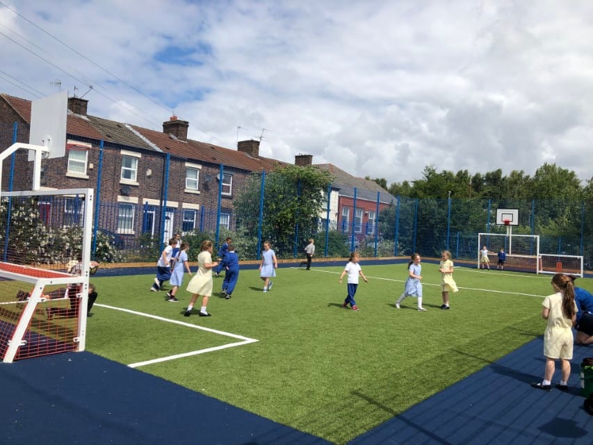 A large class of children are playing on a synthetic grass pitch, which has two goals set up on either end, playing a game of football.