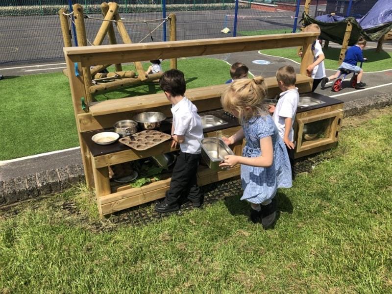 3 children playing at a mud kitchen, one girl wearing a blue summer dress and two boys wearing white shirts whilst other children play in the background. 
