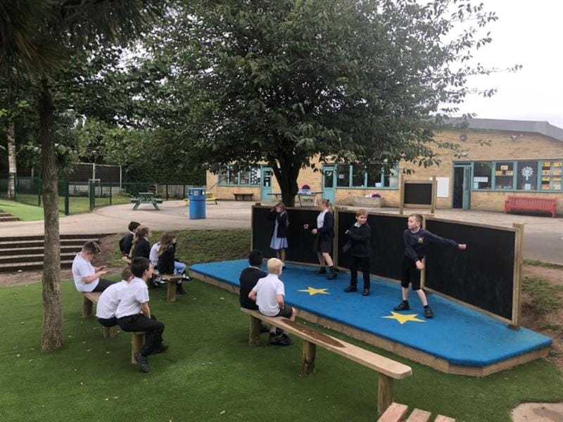children stand on the blue surfaced performance stage dancing and singing for their classmates who sit on robinia perch benches watching
