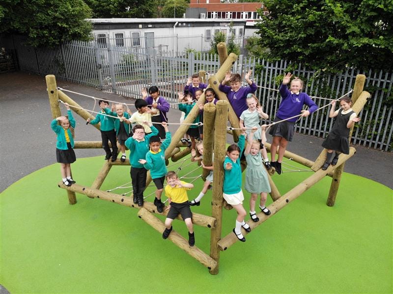 Children in purple, green and yellow school uniforms climb on one of of climbing frames whilst looking and waving up at the drone camera