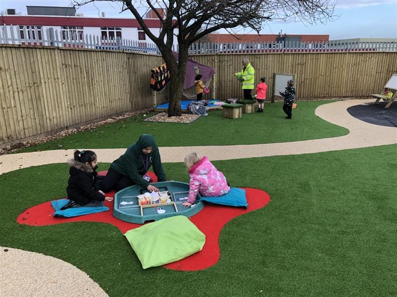 children sit on the floor with a teacher and play with items in a tuff spot tray