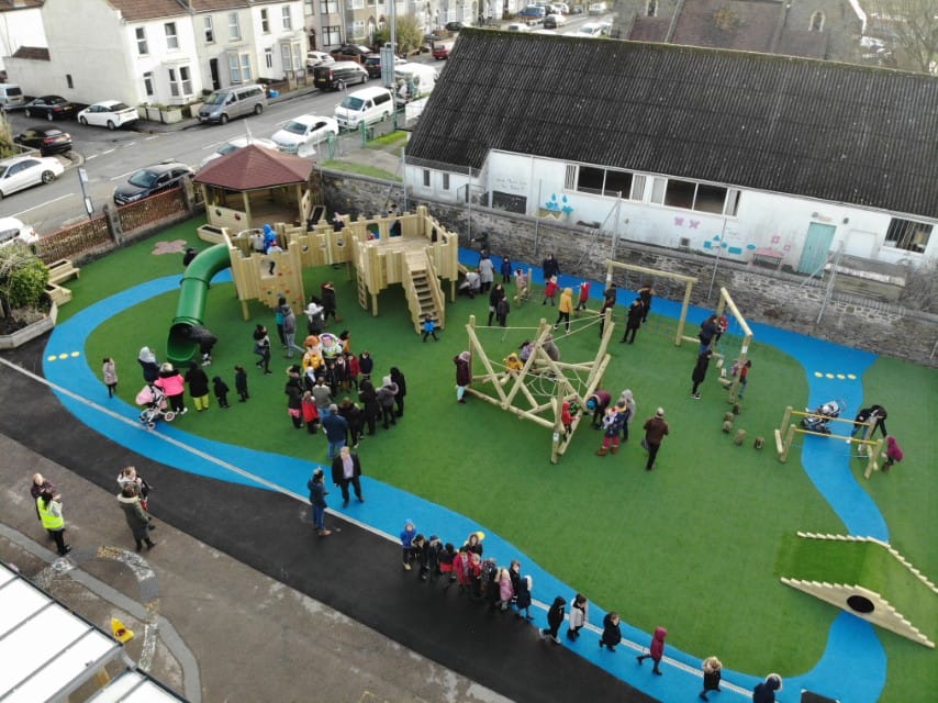 A large group of children are playing on an artificial grass surface with a variety of active play equipment.