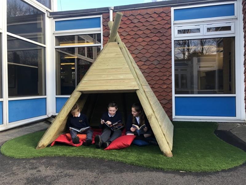 3 children sat on red cushions inside of a wigwam reading storybooks, wigwam has been installed onto artificial grass in front of the school building. 
