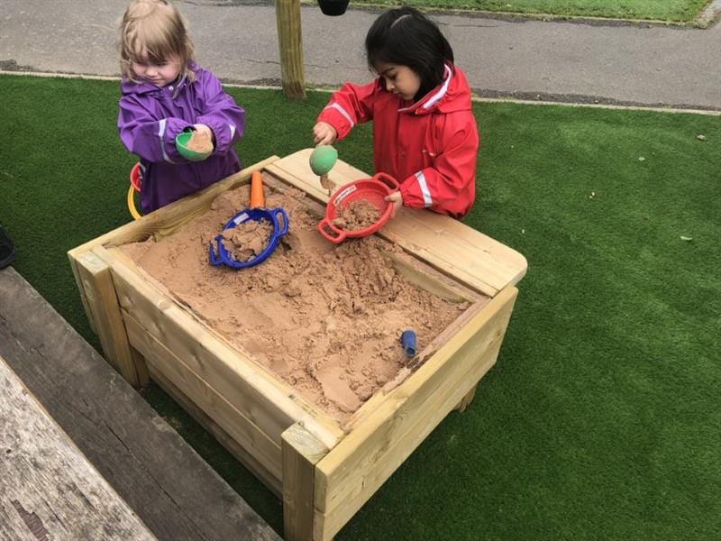 2 girls, one wearing a purple coat and one wearing a red coat are playing with sand in a sandbox. The sandbox has been installed onto artificial grass next to a school path. 