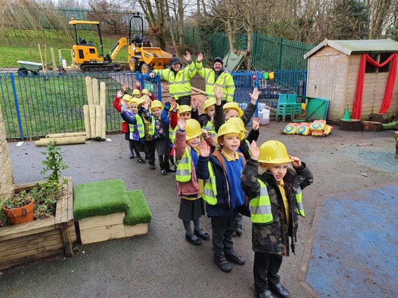 2 installers wearing fluorescent jackets and black hats stood behind 12 children who are stood in a line wearing hi-vis jackets and yellow builders hats, all waving to the camera. Behind them is two yellow diggers next to green fencing. 