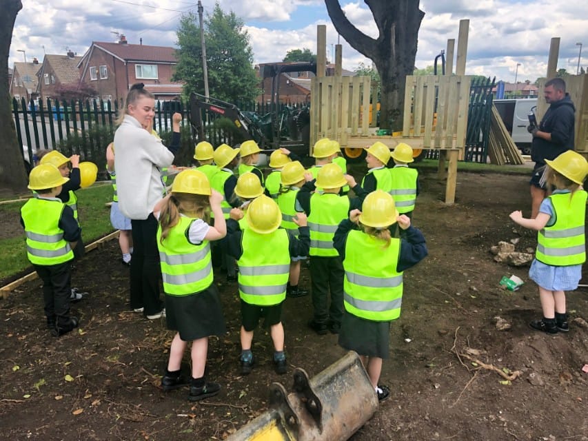 A group of children are stood outside wearing construction outfits as they watch their playground be built,