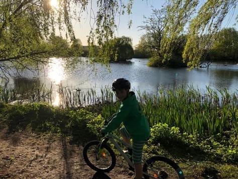 A little boy is sat on his bike and is looking at the camera as a lake is behind him.