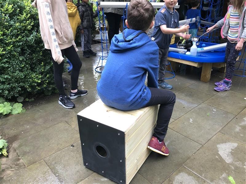 A boy is sat on a drum seat facing away from the camera and is looking at other children close by. On the back of the drum seat is a hole which is wear the sound is produced from.