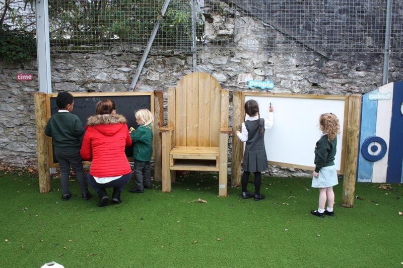 2 children stood at a chalkboard and 2 children stood at a whiteboard with one teacher in a red coat supervising. There is a storytelling chair in the middle of the chalkboard and whiteboard which have been installed onto artificial grass. 
