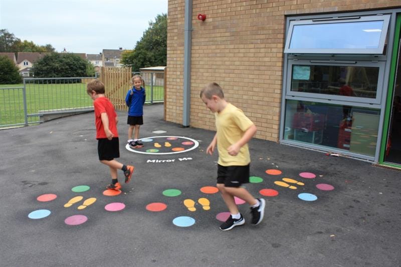 Three children, one wearing a yellow top, one wearing a red top, one wearing a blue top are playing on playground markings that have been put on the playground in front of the school building. 