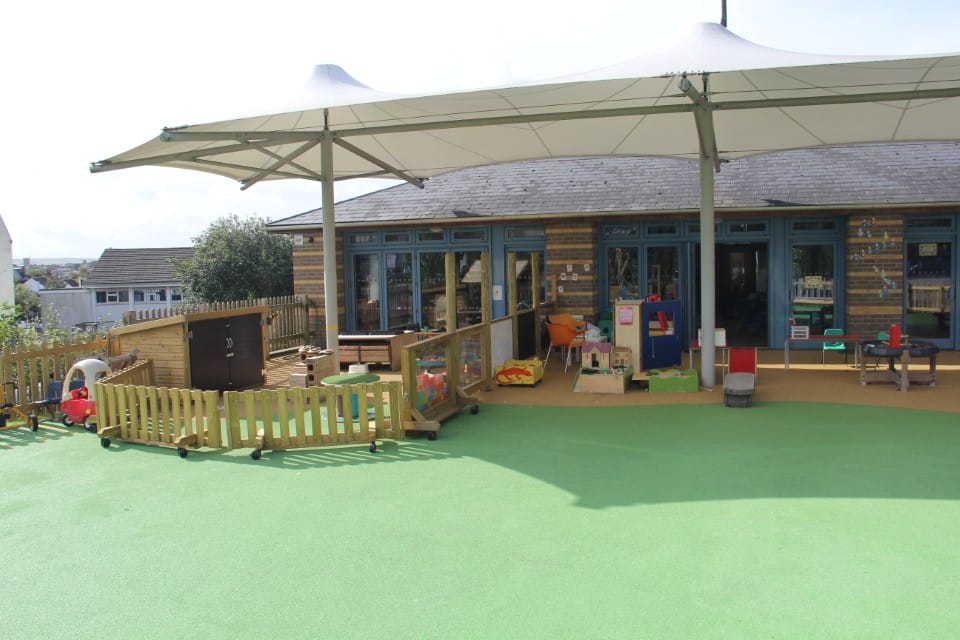 A green wetpour floor that is near a school building. The school has a canopy attached to it, with the wetpour flooring being underneath it. The majority of the wetpour is green, with a slight bit being orange.