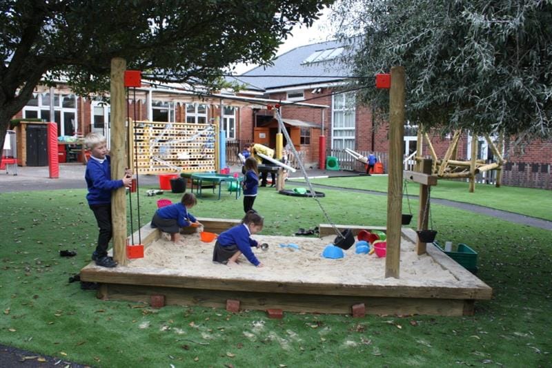 3 children wearing blue school jumpers are playing in a sandpit that has a rope and pulley, it has been installed in between two large trees in front of a waterwall. 
