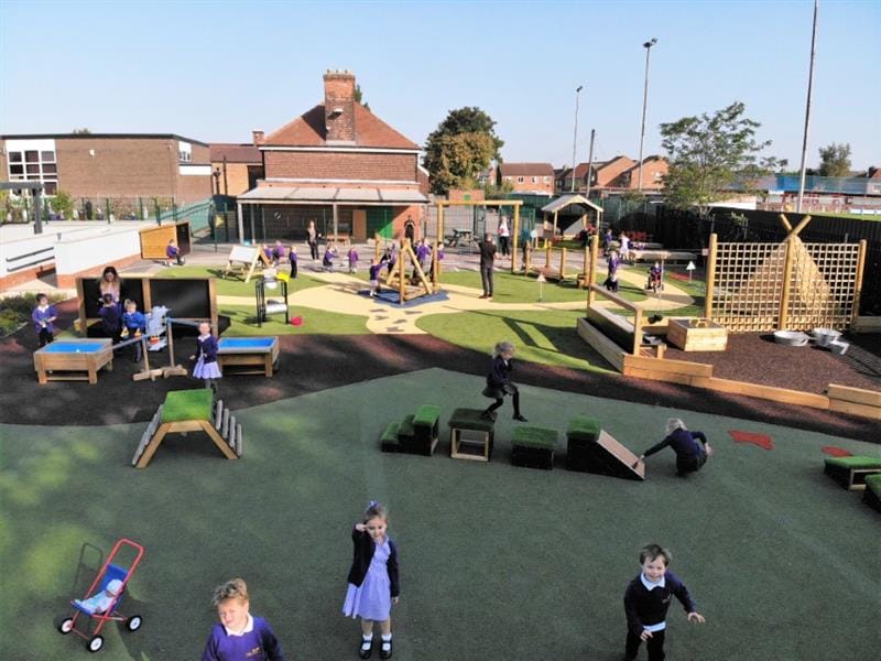 A huge playground development including a messy play area, artificial grass, a climbing frame, a giant playhouse and a wigwam. There are lots of children playing on the playground in the sunshine. 