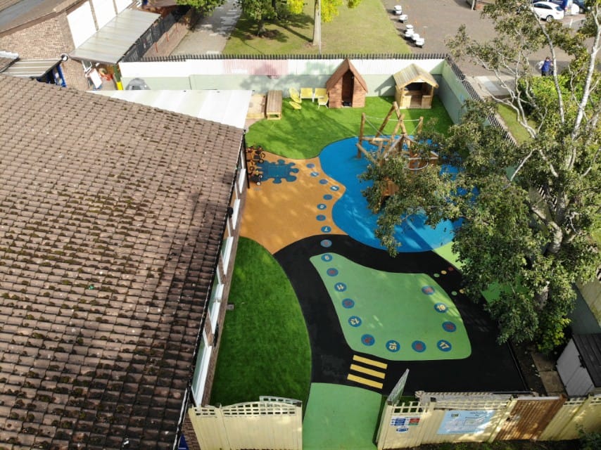 Drone shot of a playground with different wetpour sections. There's a green section with a road, a blue section with a climbing frame and an orange section which has a wetpour graphic of a splash. There's a wetpour graphic of a number snaking going from the green section to the orange section.