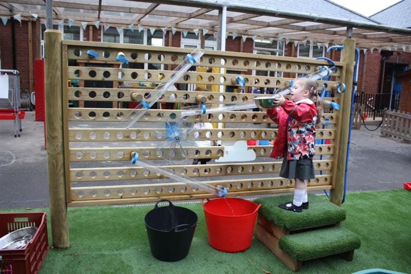 a little girl stands on the artificial grass topped steps to reach the top of the tubes on the water wall as she pours water down the tubes and into the buckets below