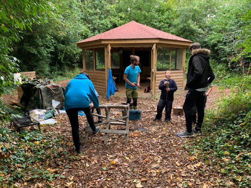 A group of people are stood out the front of a 5M Hexagonal Gazebo, made by Pentagon Play. The gazebo has been installed in a forest and is surrounded by trees and foilage.