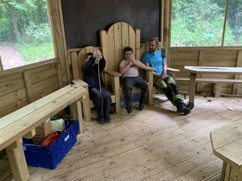 2 boys sat on storytelling chairs inside of a hexagonal gazebo with one installer wearing a bright blue top also sat on a storytelling chair talking to the two boys. 