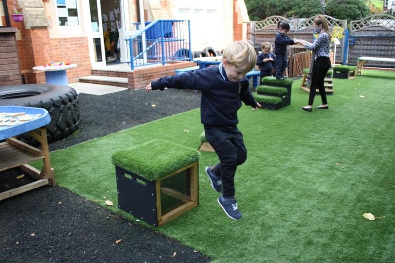 One boy with blonde hair is jumping off a get, set go! block that has been placed onto artificial grass whilst one girl stands on another block holding hands with a teacher. 