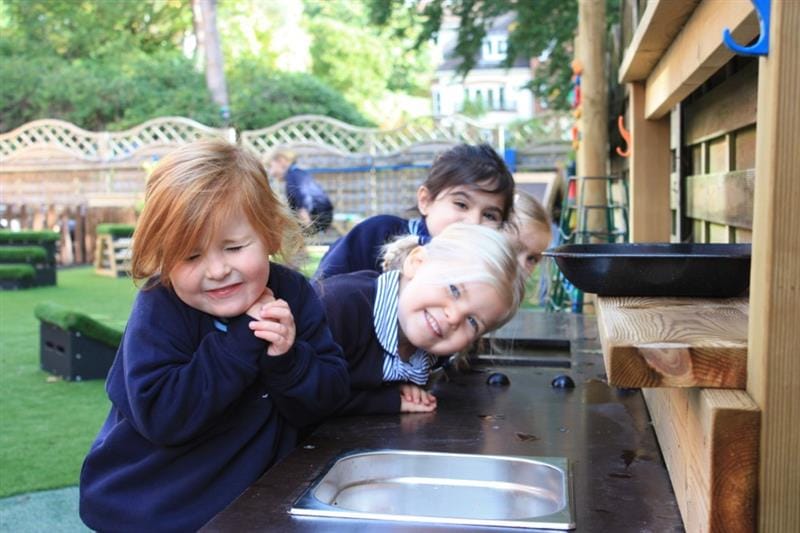 three children stand by the mud kitchen and smile at the camera