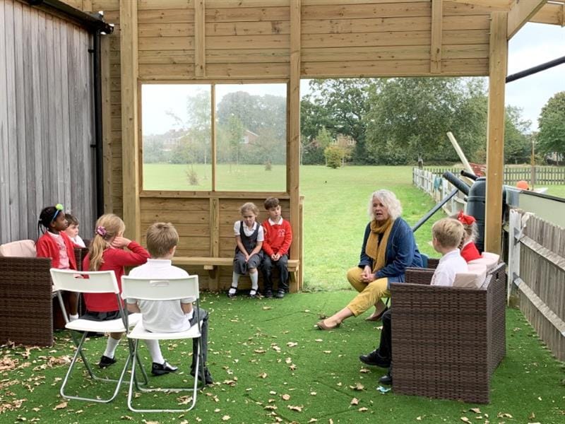 children sit in a circle in an outdoor classroom with a teacher and they are talking
