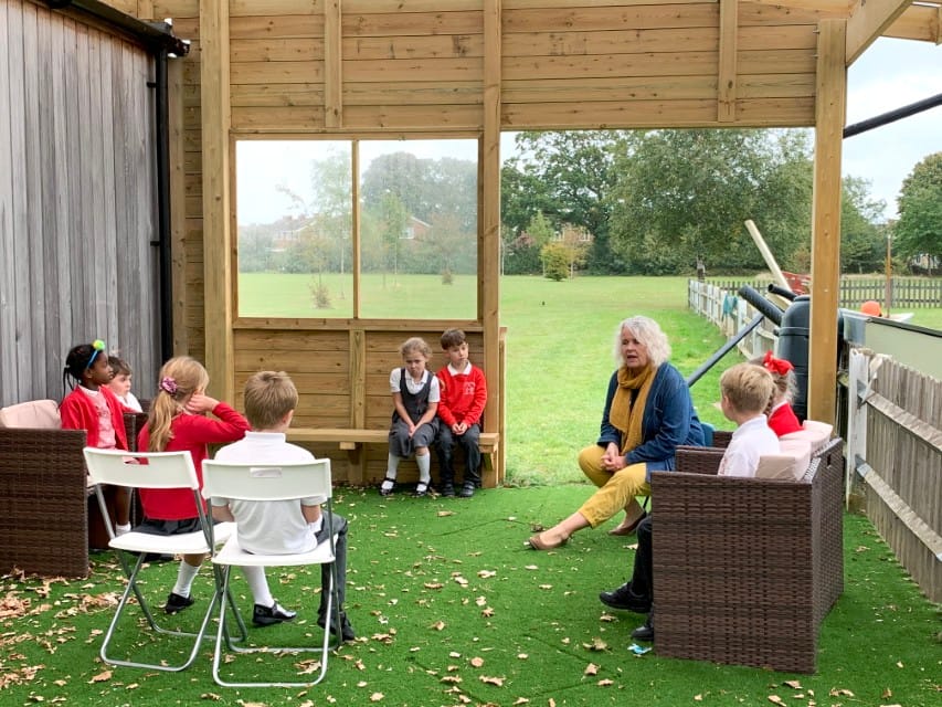 A teacher is sat outside with a group of children inside an outdoor classroom. They are sat on chairs which have been placed on artificial grass surfacing.
