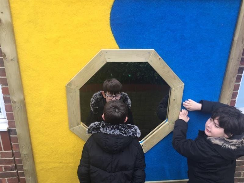 Two children engaging their senses on a large sensory panel. One is looking at their reflection in the mirror whilst the other touches the blue saferturf surfacing