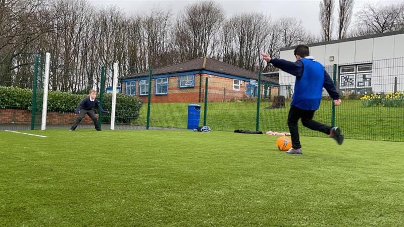 One boy about to kick a bright orange football aiming for a goal on a MUGA pitch whilst another boy wearing school uniform stands in the goal net. 