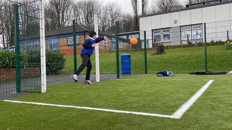 One boy wearing school uniform and blue vest playing on a MUGA pitch with a bright orange football, installed in front of school building.