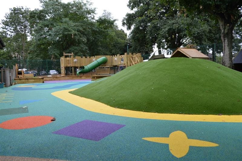 Blue playground surfacing with red, purple, yellow and green shapes on with a green mound covered in artificial grass in the middle. There is a large castle play frame in the background with a huge green slide. 