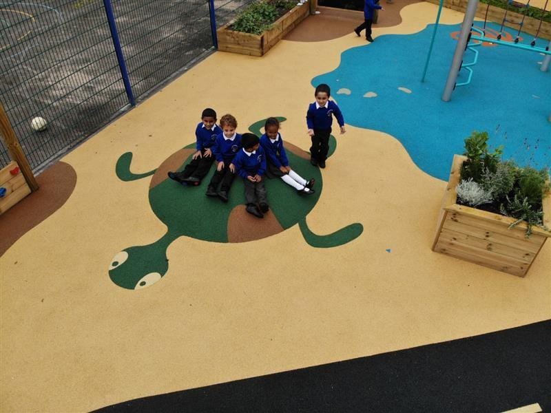 4 children wearing blue school jumpers sat on a turtle feature surface mound smiling at the camera whilst one boy stands on the edge of the mound near a square planter. 