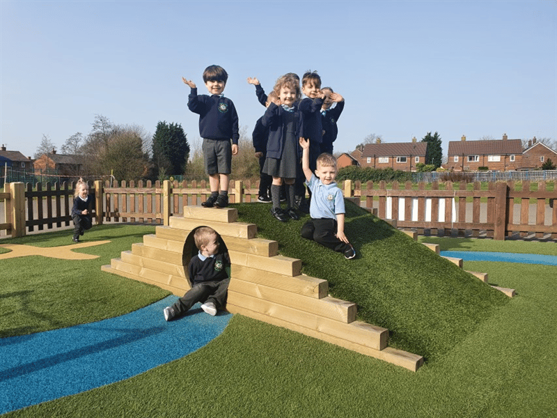 6 children stood on a climb through tunnel which has been topped with artificial grass, waving and smiling to the camera whilst one child is running towards the hill and one child sat inside of the tunnel. 
