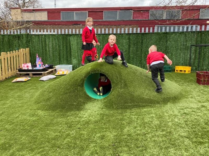 4 children playing on top of a climb through tunnel that has been topped with artificial grass that has been installed in front of the school building whilst one child sits inside of the tunnel.