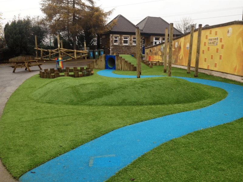 Playground surface mound placed in the middle of school playground which is surrounded by a blue path with a climb through tunnel in the background.