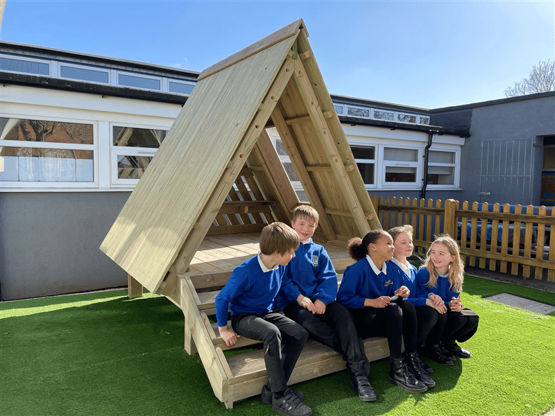 5 children in blue school jumpers sat on the steps of a forest floor learning den socialising