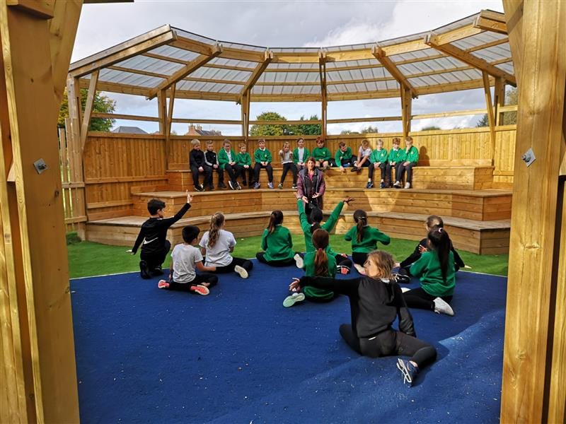 a playground ampitheatre with the performance stage in front as people sit with people watching their peers in green school uniform performing