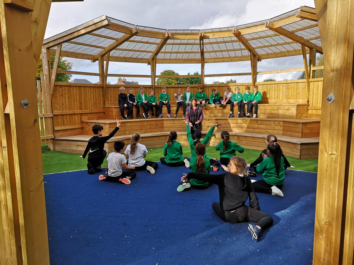 A group of children are sat on a blue stage as a teacher looks out to them. Another group of children are sat on seating facing the stage and are watching.