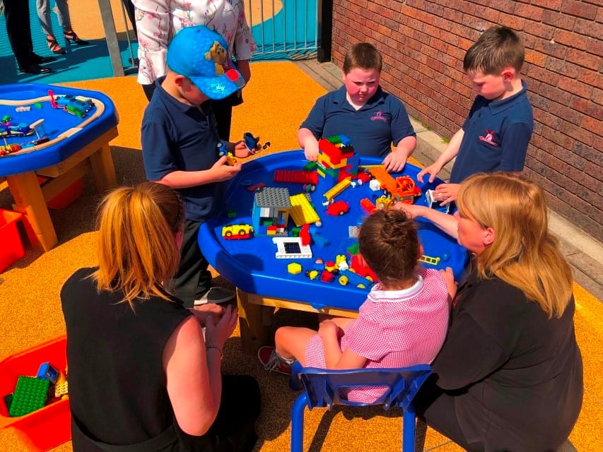 A group of children are playing with toys that are inside a tuff tray. Two teachers are crouching next to a little girl and are playing with the group.