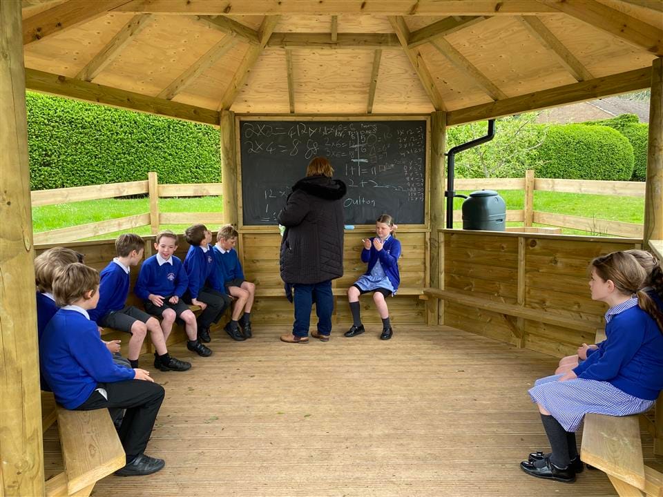 A teacher is writing multiple maths questions on a chalkboard, as her class is watching her write on the board.