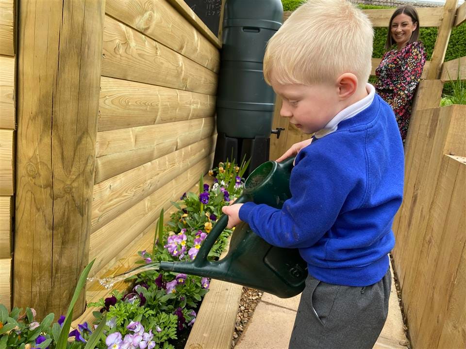 A little boy is watering flowers that have been planted in a wooden planter. A teacher is behind him, smiling.
