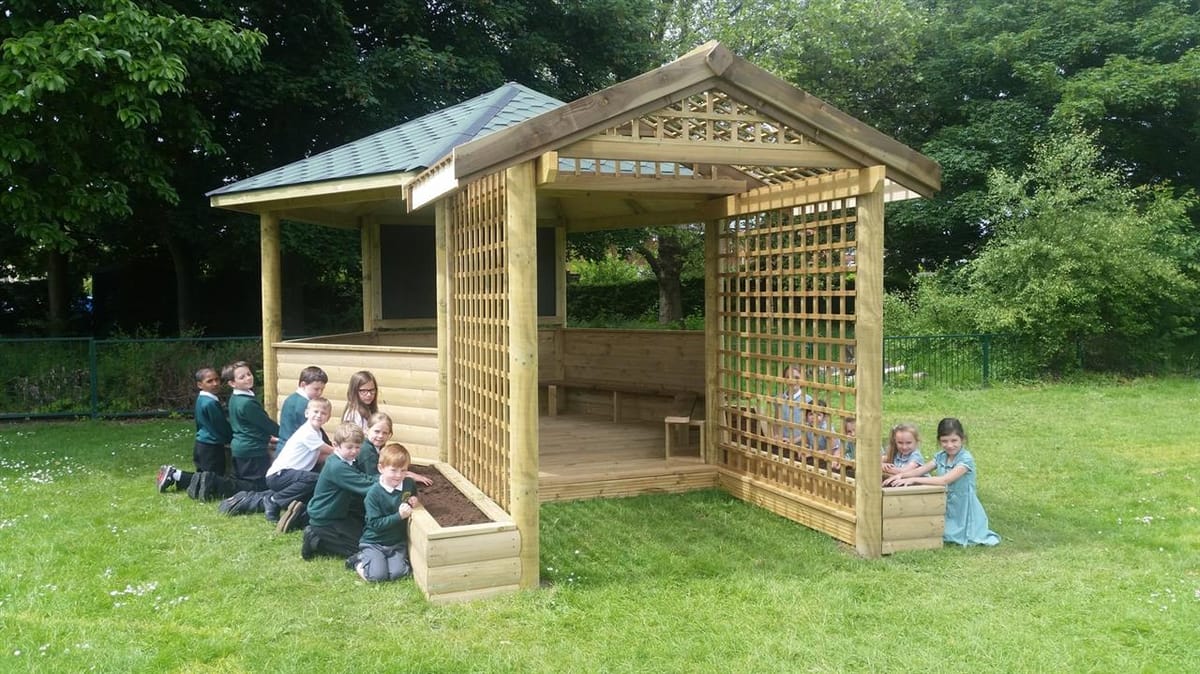 A large group of children are kneeling beside the planters that go around the outdoor gazebo. They are all looking at the camera and smiling.