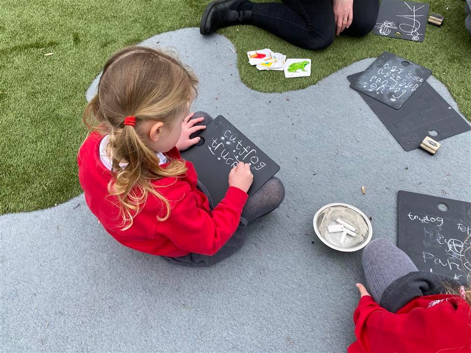 A little girl is writing on a small chalkboard that she has in her lap. She is writing different words on the board as she draws lines across the board. A teacher and another student are sat next to her.
